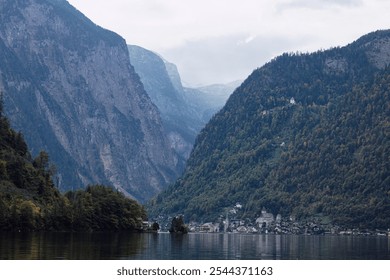 This photo depicts a serene lakeside village nestled between towering mountains, with calm waters reflecting the landscape. The scene is tranquil and surrounded by dense forests and steep cliffs. - Powered by Shutterstock