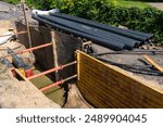 This photo depicts a trench at a construction site, secured with shoring to prevent collapse. Several large pipes are stacked nearby, ready for installation. The trench is partially filled with water