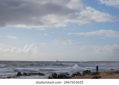 In this photo, a fisherman is depicted catching fish while standing on the shore of the ocean. He stands at the edge of a rocky coastline, holding a long fishing rod, its tip dipping into the churning - Powered by Shutterstock