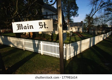 This Is A Photo Of A Typical Suburban House On Main Street, USA. There Is A White Picket Fence On A Shaded, Tree Lined Street With A Green Lawn. The Corner Has Sign Saying Main Street & Front Street.