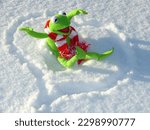 In this photo you can see the popular Muppet character Kermit sitting in the snow. There is a heart drawn around him Kermit is fully visible wearing a scarf Kermit is pleased smiling and looking ahead