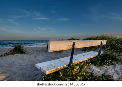 This photograph captures a white wooden bench positioned on a sandy beach, overlooking a calm ocean under a clear evening sky. - Powered by Shutterstock