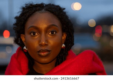 This photograph features a front-facing close-up of a serene young woman with a gentle, engaging gaze. She is wrapped in a soft, scarlet wrap that stands out against the dimming light of a city at
