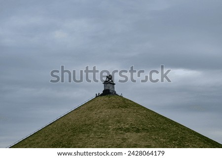 This photograph presents a somber view of visitors ascending the Lion's Mound, the prominent memorial at the site of the Battle of Waterloo. The overcast sky and the mound's steep incline lead the eye
