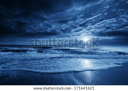 This is a photographic illustration of a beautiful midnight blue ocean moonrise along the coast with dramatic sky and rolling waves.