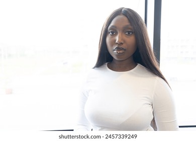 This portrait captures a young Black woman in a white long-sleeve top, posed against a softly blurred urban backdrop seen through a large window. The natural lighting highlights her composed - Powered by Shutterstock