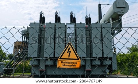 This powerful photograph features a bright yellow high voltage warning sign prominently displayed on a chain link fence in the foreground, with a large industrial transformer and its bushings visible in the background. The composition creates a strong safety message combined with technical industrial detail under natural daylight.
