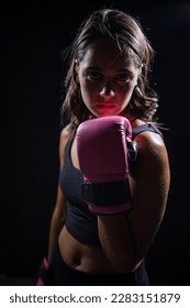 This powerful stock photo features a pretty dark-haired girl boxing a punching bag in a dark and moody setting. She is wearing boxing gloves and is fully focused on her training or exercise routine. 