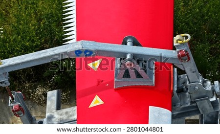 This precise top-down aerial photograph provides a detailed close-up view of the specialized lifting and attachment rig connected to a large modern wind turbine rotor blade during the construction phase. The image clearly shows the heavy-duty metal framework, chains, hooks, and safety mechanisms securely fastened to the red and white painted blade section with serrated trailing edge.