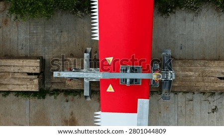 This precise top-down aerial photograph provides a detailed view of three large modern wind turbine rotor blades with distinctive red and white coloring and serrated trailing edges, laid out on a construction site within a vibrant rural agricultural landscape. The high-resolution drone perspective clearly showcases the impressive length, technical design, and aerodynamic features of the blades, positioned on a prepared platform surrounded by green vegetation and expansive crop fields.
