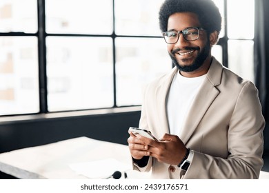 This professional using his smartphone standing in a stylish office setting. The large windows behind him fill the room with light. African-American male employee texting online on the mobile phone - Powered by Shutterstock