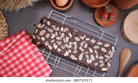 This is a rectangular-shaped loaf of chocolate-colored, fudgy brownie, covered entirely on its top with slivered almonds in a neat, overlapping pattern.  The brownie is placed directly on a metal wire cooling rack, which is sitting on a gray, slate-like countertop.  In the upper right corner, there are three whole red strawberries with green leafy tops. In the upper center, there is a brown-shelled egg resting in a small wooden cup. To the left of the cooling rack is a folded red and white checkered cloth, and in the bottom right corner is a natural wood spoon with a round head and straight handle. The image is taken from a top-down perspective, looking directly down at the brownie and the surrounding objects.