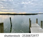 This serene image captures a calm waterfront at sunset, with soft ripples reflecting the pastel sky. Wooden posts from an old dock stand quietly in the still water, bordered by lush greenery on the ho