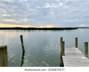 This serene image captures a calm waterfront at sunset, with soft ripples reflecting the pastel sky. Wooden posts from an old dock stand quietly in the still water, bordered by lush greenery on the ho - Powered by Shutterstock