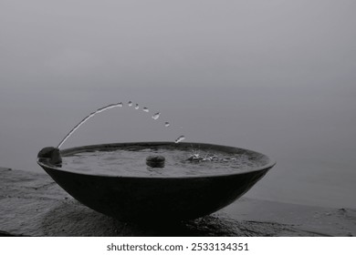 This serene image captures a minimalist water fountain with droplets arching from a metal bowl. Set against a foggy lake, it evokes calmness, ideal for wellness, spa, or zen-inspired projects. - Powered by Shutterstock