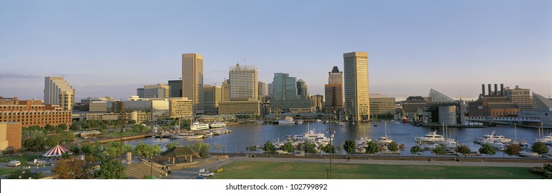 This is the skyline of Baltimore in daylight showing the inner harbor and city lights. There are boats moored in the harbor