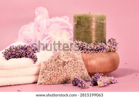 This spa essentials setup showcases a stack of white towels, a green candle, a wooden bowl with lavender, a sheer sachet of dried lavender, and a pink bath pouf, all on a soft pink surface.