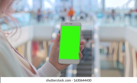 This stock photo features a woman holding her smartphone with green screen as she stands in a shopping mall