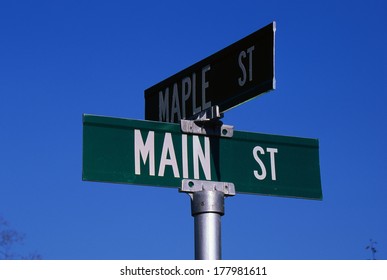 This Is A Street Sign Labeling The Corner Of Main Street & Maple Street. The Sign Is Green With White Lettering Against A Blue Sky.