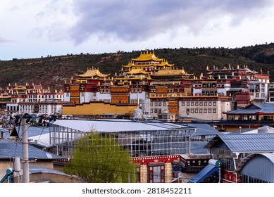This striking image presents a panoramic view of a grand Tibetan Buddhist monastery in Yunnan, China, set against a dark, rugged hillside beneath a moody, overcast sky. The magnificent temple complex features traditional architecture with vibrant red and white walls, crowned by gleaming, ornate golden roofs that immediately draw the eye. In the foreground, contemporary town infrastructure, including utility lines and a modern glass-roofed structure, creates a fascinating contrast between ancient spiritual heritage and ongoing urban development. The sprawling scale of the religious landmark, reminiscent of the famous Songzanlin Monastery in Shangri-La, dominates the landscape, exuding a sense of peace and historical grandeur. This captivating, highly detailed photograph is perfect for travel promotions, cultural documentaries, and editorial projects highlighting the intersection of ancient religious traditions and modern daily life in highland Asia.