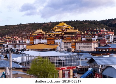 This striking image presents a panoramic view of a grand Tibetan Buddhist monastery in Yunnan, China, set against a dark, rugged hillside beneath a moody, overcast sky. The magnificent temple complex features traditional architecture with vibrant red and white walls, crowned by gleaming, ornate golden roofs that immediately draw the eye. In the foreground, contemporary town infrastructure, including utility lines and a modern glass-roofed structure, creates a fascinating contrast between ancient spiritual heritage and ongoing urban development. The sprawling scale of the religious landmark, reminiscent of the famous Songzanlin Monastery in Shangri-La, dominates the landscape, exuding a sense of peace and historical grandeur. This captivating, highly detailed photograph is perfect for travel promotions, cultural documentaries, and editorial projects highlighting the intersection of ancient religious traditions and modern daily life in highland Asia.