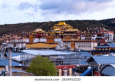This striking image presents a panoramic view of a grand Tibetan Buddhist monastery in Yunnan, China, set against a dark, rugged hillside beneath a moody, overcast sky. The magnificent temple complex features traditional architecture with vibrant red and white walls, crowned by gleaming, ornate golden roofs that immediately draw the eye. In the foreground, contemporary town infrastructure, including utility lines and a modern glass-roofed structure, creates a fascinating contrast between ancient spiritual heritage and ongoing urban development. The sprawling scale of the religious landmark, reminiscent of the famous Songzanlin Monastery in Shangri-La, dominates the landscape, exuding a sense of peace and historical grandeur. This captivating, highly detailed photograph is perfect for travel promotions, cultural documentaries, and editorial projects highlighting the intersection of ancient religious traditions and modern daily life in highland Asia.