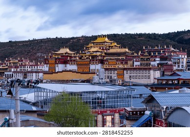 This striking image presents a panoramic view of a grand Tibetan Buddhist monastery in Yunnan, China, set against a dark, rugged hillside beneath a moody, overcast sky. The magnificent temple complex features traditional architecture with vibrant red and white walls, crowned by gleaming, ornate golden roofs that immediately draw the eye. In the foreground, contemporary town infrastructure, including utility lines and a modern glass-roofed structure, creates a fascinating contrast between ancient spiritual heritage and ongoing urban development. The sprawling scale of the religious landmark, reminiscent of the famous Songzanlin Monastery in Shangri-La, dominates the landscape, exuding a sense of peace and historical grandeur. This captivating, highly detailed photograph is perfect for travel promotions, cultural documentaries, and editorial projects highlighting the intersection of ancient religious traditions and modern daily life in highland Asia.