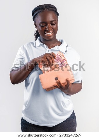 This studio portrait features a young woman with a warm, joyful expression, captured against a clean, neutral white background. She has deep-toned skin and her hair is neatly styled in thick, dark cornrow braids that transition into a ponytail at the back. She is dressed casually in a crisp, short-sleeved white polo shirt with thin dark piping on the collar and sleeves, paired with dark denim jeans.

The central focus of the image is the lady's interaction with a peach or salmon-colored rectangular purse. She holds the purse with the left hand while the right hand is tucked inside, pulling out or organizing several banknotes. The visible currency consists of 200 Ghanaian Cedi notes, identifiable by their distinct red and orange color palette and numerical markings. Her gaze is directed downward toward the money, and she wears a bright, satisfied smile that suggests financial success, savings, or a positive transaction. A simple gold bracelet adorns her right wrist, adding a subtle touch of elegance to her functional, everyday look. The lighting is soft and even, highlighting features and the texture of the currency.