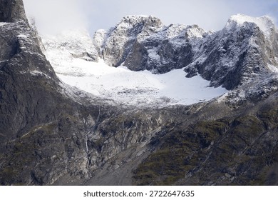 This stunning drone image captures a high-altitude mountain landscape, showcasing rugged peaks dusted with snow under soft, diffuse lighting. The photograph, taken from an aerial perspective, highlights the dramatic contrasts between dark rocky cliffs and the bright white snowfields, set against a backdrop of cloudy skies.