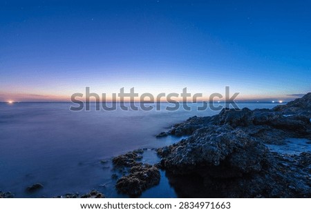This stunning image captures the tranquil beauty of a coastal landscape at twilight. The scene features rugged rocks in the foreground, partially covered with seaweed, gently meeting the calm ocean waters. The long exposure technique creates a smooth, mist-like effect on the sea, enhancing the serene atmosphere. The horizon glows with a soft gradient of warm orange and yellow tones, fading into deep blue as night approaches. Distant lights from boats or ships shimmer across the horizon, adding subtle points of interest. Above, faint stars begin to appear in the clear evening sky. The composition evokes a sense of peace, solitude, and natural wonder, making it ideal for use in travel, nature, or landscape-themed projects. This photo perfectly illustrates the harmony between land, sea, and sky during the magical transition from day to night.