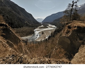 This stunning view, taken from a dramatic foreground cliff, captures the Rhine River meandering through a deep, mountainous valley. The river flows across a wide, sandy floodplain, bordered by steep, wooded slopes characteristic of the Swiss Alps. The scene captures a tranquil spring day, emphasizing the contrast between the rushing water, the dramatic cliffs, and the lush alpine backdrop. The location is in the Rheintal, Switzerland.