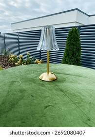 This stylish table lamp features a striking gold base and elegant crystallike shade, placed on a luxurious green velvet table in an inviting outdoor setting, surrounded by vibrant foliage