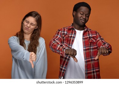 This Sucks. Portrait Of Emotional Young Interracial Couple African Male And Caucasian Woman Showing Thumbs Down Gesture, Disgusted With Bad Food Or Stink, Suppressing Vomiting. Dislike And Disgust