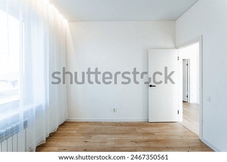 This sunlit, spacious room features white walls, sheer curtains, and wooden flooring. An open door connects it to another bright space. Ideal for real estate listings and design inspiration