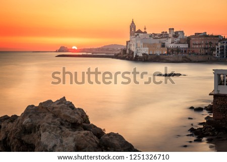 This is a sunset view with rocks on the foreground, the mediterranean sea blurried by a slow shutter speed and the town of sitges in the background.