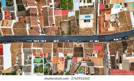 This top-down aerial view captures the geometry of a Mediterranean village street. White houses with red terracotta roofs surround a central road with parked cars, revealing private courtyards and a small swimming pool from a drone perspective.