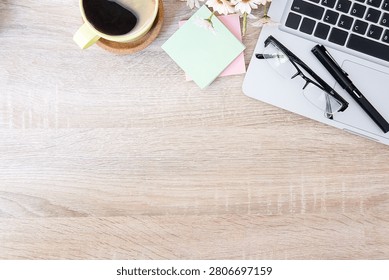 This top-down shot of a bright, light wood desk shows a neat work setup pushed toward the top edge. On the right, a silver laptop serves as a resting spot for a pair of black-rimmed reading glasses and a black fountain pen. Right next to it sits a small stack of pink and mint green sticky notes, tucked under a few delicate white daisies. Over in the upper left corner, a yellow mug on a cork coaster is filled with black coffee. The whole bottom half of the image is wide-open, natural wood grain surface, leaving plenty of clean space for text or layout designs.