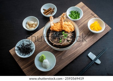 This is a top-down, slightly angled shot of a bowl of Japanese soba noodles with two pieces of shrimp tempura on top, surrounded by small bowls of various condiments