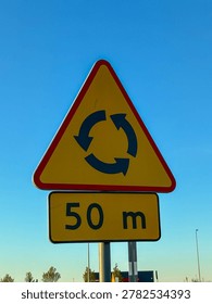 This triangular road sign featuring a roundabout symbol and distance plate warns motorists of an upcoming traffic circle to promote road safety and effective urban transit flow
