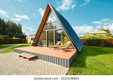 This unique A-frame house features large glass windows and a wooden deck, surrounded by green grass and trees under a bright blue sky. Two lounge chairs sit invitingly on the deck. - Powered by Shutterstock