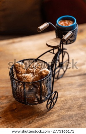 This unique image showcases a creative food presentation featuring crispy fried snacks served in a miniature wire bicycle basket. A small blue bowl of red dipping sauce rests on the handlebars, all on a warm wooden surface.