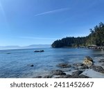 This Vancouver Island landscape is a serene and picturesque view of the ocean, with rocky shorelines, green pine trees, and distant snow-capped mountains under a clear blue sky.