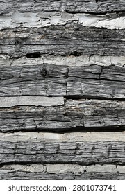This is a vertical close up of weathered flat log cabin logs with natural patina, cracked uneven wood, cut marks, and porous chinking. The chinking is chipped with some missing areas, and a few rusty nails are visible in the logs.