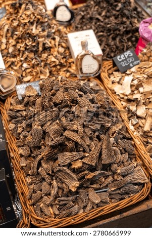 This vertical image captures the intricate textures of dried morel mushrooms piled high in a rustic wicker basket. In the blurred background, other varieties like Ceps (Porcini) and specialty heart-shaped oil bottles are visible, typical of a high-end European food market.