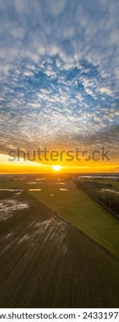 This vertical portrait captures the grandeur of a sunset descending over farmlands. The sky, filled with a blanket of textured clouds, is aflame with the golden hues of the setting sun. Below, the