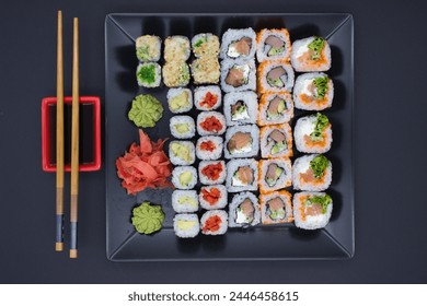 This vibrant image captures a variety of sushi rolls neatly displayed on a large black plate, accompanied by bamboo chopsticks, a red soy sauce dish, wasabi, and pickled ginger. The assortment include