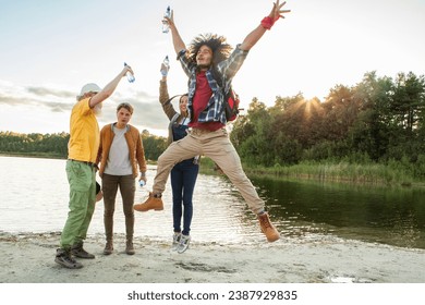 In this vibrant photo, a diverse group of millennial friends celebrates their arrival at a forest lake. Water bottles are joyfully raised in the air, capturing a moment of triumph. The excitement - Powered by Shutterstock