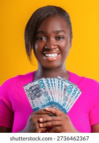 This vibrant studio portrait features a smiling young woman with a warm, joyful expression, set against a solid, bright yellow background that makes the colors pop. She is positioned centrally, facing the camera, and holds a fanned-out stack of Ghanaian currency—specifically 100 Cedi notes—with both hands. The woman has dark skin and styled dark hair with a side-swept fringe. She is wearing a bright magenta or hot pink short-sleeved top that contrasts sharply with the yellow backdrop.

The currency notes are teal and blue, featuring the 