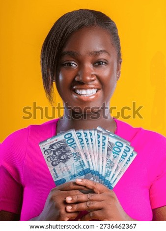 This vibrant studio portrait features a smiling young woman with a warm, joyful expression, set against a solid, bright yellow background that makes the colors pop. She is positioned centrally, facing the camera, and holds a fanned-out stack of Ghanaian currency—specifically 100 Cedi notes—with both hands. The woman has dark skin and styled dark hair with a side-swept fringe. She is wearing a bright magenta or hot pink short-sleeved top that contrasts sharply with the yellow backdrop.

The currency notes are teal and blue, featuring the 