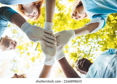 In this view from below, a group of unrecognizable volunteers stand in a circle and fold their arms, are casually dressed and standing in the grass outdoors.